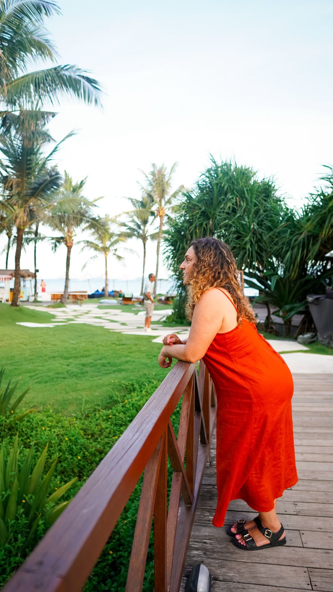 Woman in an orange dress resting on a timber railing among palm trees and lawn